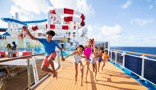 A group of 6 young children holding hands and playing on deck of a Carnival cruise ship. On deck playground and slide in background with view of ocean.